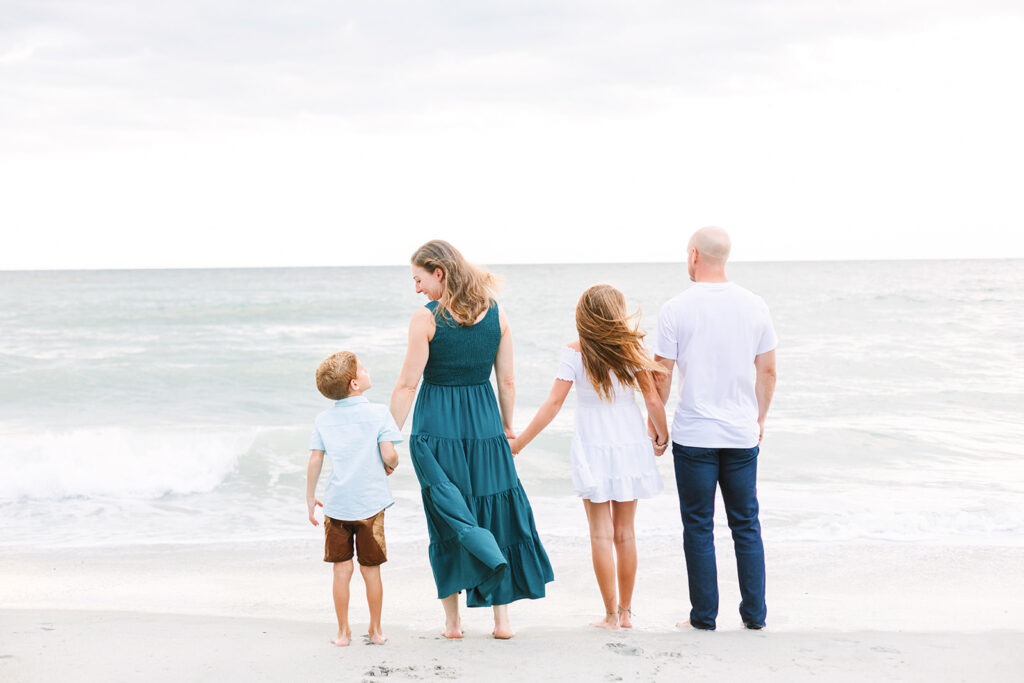 Family walking on the sand coastline facing away from the camera in neutral tones with pops of a blue and green color, JCrane Photography, Best family photographer in North Carolina, Top-rated family photographer North Carolina, Outdoor Family Photographer Near Me