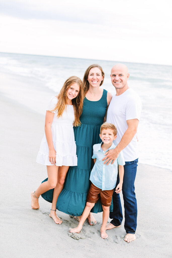 Family standing on beach facing the camera in neutral tones with pops of a blue and green color, JCrane Photography, Best family photographer in North Carolina, Top-rated family photographer North Carolina, Outdoor Family Photographer Near Me