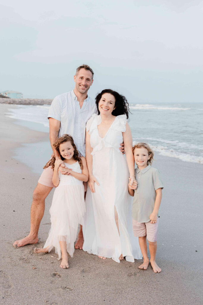 Family standing on the beach in soft neutral coastal tone clothes looking at the camera, JCrane Photography, Best family photographer in North Carolina, Top-rated family photographer North Carolina, Outdoor Family Photographer Near Me